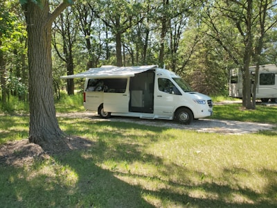 A white recreational vehicle is parked on a grassy area surrounded by a forest of tall trees. The vehicle has its side canopy extended, providing shade. Sunlight filters through the leaves, creating a dappled pattern on the ground.