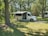 A white recreational vehicle is parked on a grassy area surrounded by a forest of tall trees. The vehicle has its side canopy extended, providing shade. Sunlight filters through the leaves, creating a dappled pattern on the ground.
