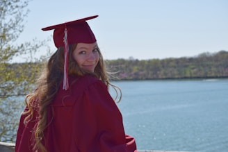 A person wearing a maroon graduation cap and gown stands by a body of water, surrounded by trees. They are facing away but look back at the camera with a smile. It's a bright, sunny day with a clear sky.