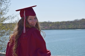 A person wearing a maroon graduation cap and gown stands by a body of water, surrounded by trees. They are facing away but look back at the camera with a smile. It's a bright, sunny day with a clear sky.