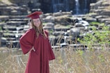 A person wearing a maroon graduation gown and cap stands against a backdrop of a natural rocky waterfall. The scene is surrounded by tall grasses and greenery, with the waterfall gently cascading in the background.