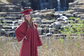 A person wearing a maroon graduation gown and cap stands against a backdrop of a natural rocky waterfall. The scene is surrounded by tall grasses and greenery, with the waterfall gently cascading in the background.