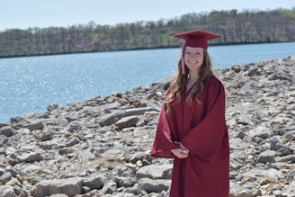 A person wearing a maroon graduation gown and cap stands on a rocky shore with a large body of water and a forested area in the background. The mood is bright due to the sunny weather and clear blue sky.