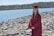 A person wearing a maroon graduation gown and cap stands on a rocky shore with a large body of water and a forested area in the background. The mood is bright due to the sunny weather and clear blue sky.