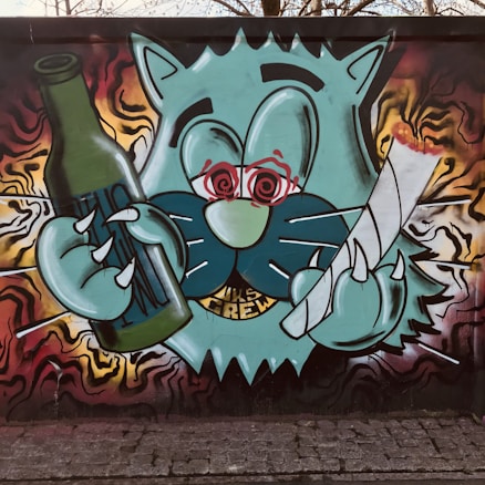 A graffiti-style mural features a whimsical, cartoon-like green cat with exaggerated features, holding a green bottle in one hand and a large feather or quill in the other. The background contains colorful abstract shapes in red, yellow, and black, adding a dynamic, energetic feel.