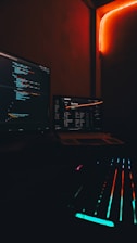 A dark, cozy room with computer screens displaying code and possibly a music playlist. Neon lights create a warm red glow, complemented by a keyboard with multicolored backlighting.