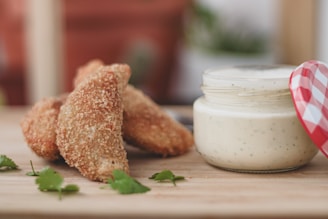 Golden brown breaded chicken wings are arranged on a wooden surface next to a jar of creamy white dip with herbs. Fresh cilantro leaves are scattered around as garnish, adding a hint of green to the composition.