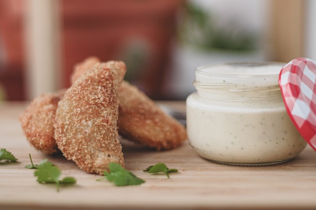 Golden brown breaded chicken wings are arranged on a wooden surface next to a jar of creamy white dip with herbs. Fresh cilantro leaves are scattered around as garnish, adding a hint of green to the composition.