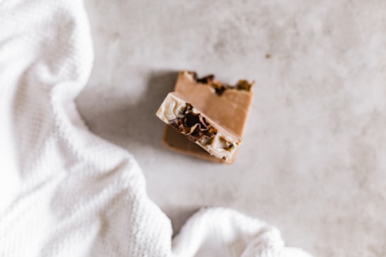 A bar of soap with a textured, creamy surface is placed on a light, soft fabric. The soap has visible brown and cream layers, giving a natural, artisanal appearance. The fabric appears to be a white, waffle-textured cloth.