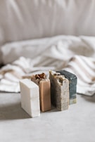 Three bars of soap in various natural colors are arranged vertically. The soaps have different textures and appear to be homemade, with one having visible seeds or grains. In the background, there's a soft, blurred fabric with stripes, adding a cozy and simple aesthetic.