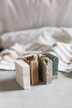 Three bars of soap in various natural colors are arranged vertically. The soaps have different textures and appear to be homemade, with one having visible seeds or grains. In the background, there's a soft, blurred fabric with stripes, adding a cozy and simple aesthetic.