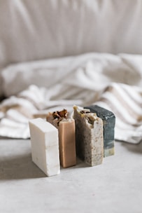 Three bars of soap in various natural colors are arranged vertically. The soaps have different textures and appear to be homemade, with one having visible seeds or grains. In the background, there's a soft, blurred fabric with stripes, adding a cozy and simple aesthetic.