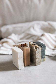 Three bars of soap in various natural colors are arranged vertically. The soaps have different textures and appear to be homemade, with one having visible seeds or grains. In the background, there's a soft, blurred fabric with stripes, adding a cozy and simple aesthetic.