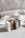 Three bars of soap in various natural colors are arranged vertically. The soaps have different textures and appear to be homemade, with one having visible seeds or grains. In the background, there's a soft, blurred fabric with stripes, adding a cozy and simple aesthetic.