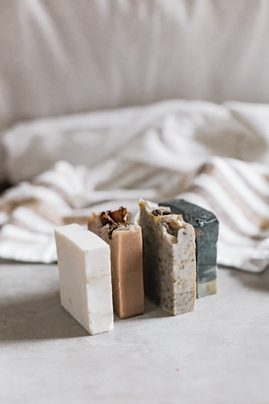 Three bars of soap in various natural colors are arranged vertically. The soaps have different textures and appear to be homemade, with one having visible seeds or grains. In the background, there's a soft, blurred fabric with stripes, adding a cozy and simple aesthetic.