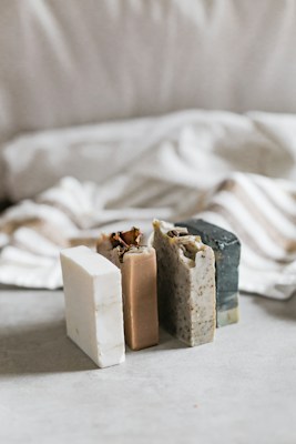 Three bars of soap in various natural colors are arranged vertically. The soaps have different textures and appear to be homemade, with one having visible seeds or grains. In the background, there's a soft, blurred fabric with stripes, adding a cozy and simple aesthetic.