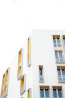 A modern building facade with clean architectural lines is featured. The walls are predominantly white with sections painted in a vibrant mustard yellow. Tall narrow windows with gray frames and small balconies are symmetrically arranged, creating an aesthetic of simplicity and elegance.