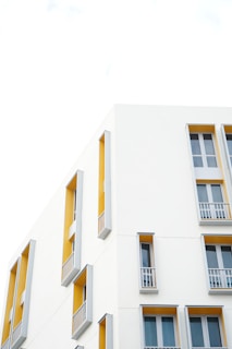 A modern building facade with clean architectural lines is featured. The walls are predominantly white with sections painted in a vibrant mustard yellow. Tall narrow windows with gray frames and small balconies are symmetrically arranged, creating an aesthetic of simplicity and elegance.
