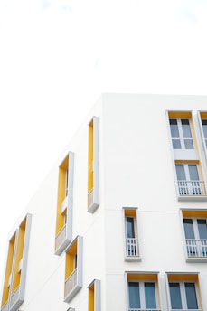 A modern building facade with clean architectural lines is featured. The walls are predominantly white with sections painted in a vibrant mustard yellow. Tall narrow windows with gray frames and small balconies are symmetrically arranged, creating an aesthetic of simplicity and elegance.
