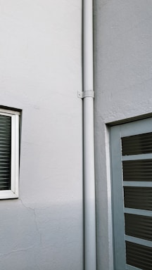 A corner of a building featuring a white drainpipe running vertically along the wall. There is a small, square window with closed blinds on the left and a door with horizontal glass panels on the right side. The walls are painted in off-white, and there are visible cracks on the surface.