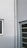 A corner of a building featuring a white drainpipe running vertically along the wall. There is a small, square window with closed blinds on the left and a door with horizontal glass panels on the right side. The walls are painted in off-white, and there are visible cracks on the surface.