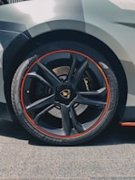 A close-up of a car wheel featuring a sleek black rim with a distinctive red trim. The wheel features a luxury brand logo in the center, and the tire has detailed tread patterns suitable for high performance. The car's bodywork is partially visible, showcasing an angular, modern design with a grey finish.