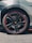 A close-up of a car wheel featuring a sleek black rim with a distinctive red trim. The wheel features a luxury brand logo in the center, and the tire has detailed tread patterns suitable for high performance. The car's bodywork is partially visible, showcasing an angular, modern design with a grey finish.