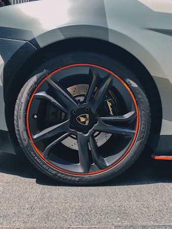 A close-up of a car wheel featuring a sleek black rim with a distinctive red trim. The wheel features a luxury brand logo in the center, and the tire has detailed tread patterns suitable for high performance. The car's bodywork is partially visible, showcasing an angular, modern design with a grey finish.