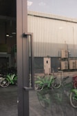 A reflective glass door with a metal handle is in the foreground, allowing a partial view of an industrial building exterior. The background reveals a metal-clad structure with several air conditioning units attached. Green bicycles are parked inside a sheltered area, partially visible through the door's reflection.