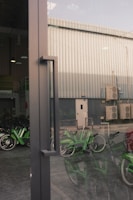 A reflective glass door with a metal handle is in the foreground, allowing a partial view of an industrial building exterior. The background reveals a metal-clad structure with several air conditioning units attached. Green bicycles are parked inside a sheltered area, partially visible through the door's reflection.