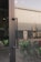 A reflective glass door with a metal handle is in the foreground, allowing a partial view of an industrial building exterior. The background reveals a metal-clad structure with several air conditioning units attached. Green bicycles are parked inside a sheltered area, partially visible through the door's reflection.