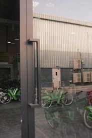 A reflective glass door with a metal handle is in the foreground, allowing a partial view of an industrial building exterior. The background reveals a metal-clad structure with several air conditioning units attached. Green bicycles are parked inside a sheltered area, partially visible through the door's reflection.
