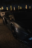 A sleek, dark-colored luxury sports car is parked on a cobblestone surface at night. Illuminated palm trees are visible in the background, casting golden light. Another vehicle, possibly an SUV, is parked nearby.