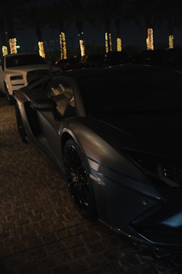 A sleek, dark-colored luxury sports car is parked on a cobblestone surface at night. Illuminated palm trees are visible in the background, casting golden light. Another vehicle, possibly an SUV, is parked nearby.