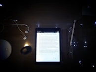 A dimly lit desk featuring a tablet displaying a document surrounded by several objects. The objects include a pair of wired earphones to the left, a wireless mouse, a stylus pen to the right, and a circular white object, possibly a wireless charging station.