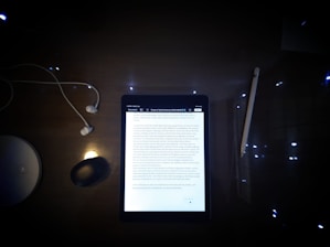 A dimly lit desk featuring a tablet displaying a document surrounded by several objects. The objects include a pair of wired earphones to the left, a wireless mouse, a stylus pen to the right, and a circular white object, possibly a wireless charging station.