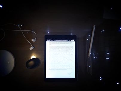 A dimly lit desk featuring a tablet displaying a document surrounded by several objects. The objects include a pair of wired earphones to the left, a wireless mouse, a stylus pen to the right, and a circular white object, possibly a wireless charging station.