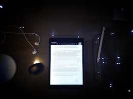 A dimly lit desk featuring a tablet displaying a document surrounded by several objects. The objects include a pair of wired earphones to the left, a wireless mouse, a stylus pen to the right, and a circular white object, possibly a wireless charging station.