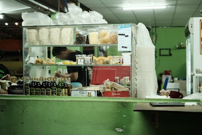 A countertop with a variety of packaged food items is displayed, including bags of dried snacks, bottles of sauces, and small packets. Behind the counter, a person is preparing something, partially visible through the glass. The setting appears to be a small food stall or shop with a green color scheme on the walls and counter.