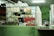 A countertop with a variety of packaged food items is displayed, including bags of dried snacks, bottles of sauces, and small packets. Behind the counter, a person is preparing something, partially visible through the glass. The setting appears to be a small food stall or shop with a green color scheme on the walls and counter.