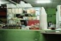 A countertop with a variety of packaged food items is displayed, including bags of dried snacks, bottles of sauces, and small packets. Behind the counter, a person is preparing something, partially visible through the glass. The setting appears to be a small food stall or shop with a green color scheme on the walls and counter.