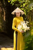 A woman wearing a traditional conical hat and a long, flowing yellow dress holds a bouquet of white lilies. She stands gracefully in a lush, green outdoor setting with sunlight filtering through the leaves.