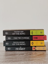 A neatly stacked set of four books in a series by J.R.R. Tolkien, including 'The Return of the King', 'The Two Towers', 'The Fellowship of the Ring', and 'The Hobbit'. The books are arranged on a wooden surface with colorful spines featuring distinct symbols and text.
