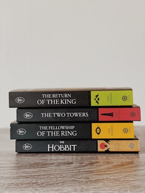 A neatly stacked set of four books in a series by J.R.R. Tolkien, including 'The Return of the King', 'The Two Towers', 'The Fellowship of the Ring', and 'The Hobbit'. The books are arranged on a wooden surface with colorful spines featuring distinct symbols and text.