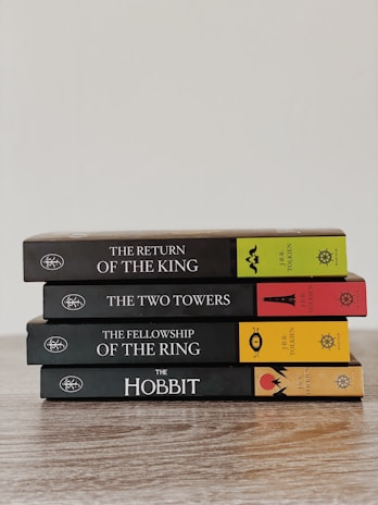 A neatly stacked set of four books in a series by J.R.R. Tolkien, including 'The Return of the King', 'The Two Towers', 'The Fellowship of the Ring', and 'The Hobbit'. The books are arranged on a wooden surface with colorful spines featuring distinct symbols and text.