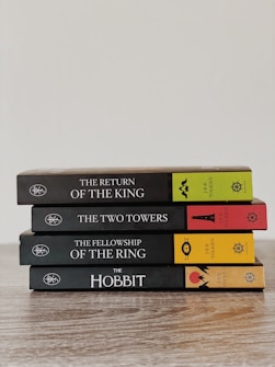 A neatly stacked set of four books in a series by J.R.R. Tolkien, including 'The Return of the King', 'The Two Towers', 'The Fellowship of the Ring', and 'The Hobbit'. The books are arranged on a wooden surface with colorful spines featuring distinct symbols and text.