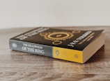 A paperback book titled 'The Fellowship of the Ring' by J.R.R. Tolkien is placed on a wooden surface. The cover features an intricate design with gold elements and text.