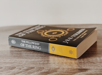 A paperback book titled 'The Fellowship of the Ring' by J.R.R. Tolkien is placed on a wooden surface. The cover features an intricate design with gold elements and text.