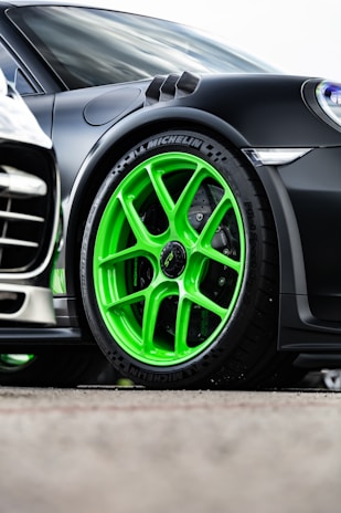 A close-up view of a sporty car featuring vibrant green alloy wheels with Michelin branding on the tires. The car has a sleek, black exterior with aerodynamic design elements. The wheel and bodywork details suggest high performance and style.