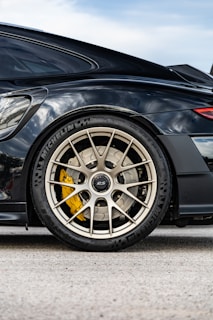 Close-up of a car's rear wheel showcasing a high-performance tire with visible branding, intricate alloy wheel design, and a bright yellow brake caliper. The car has a sleek and modern black body with reflective surfaces.